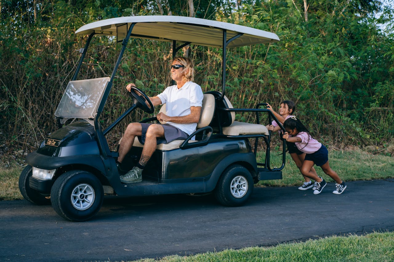 Two children playfully push a golf cart driven by an adult on a sunny day.